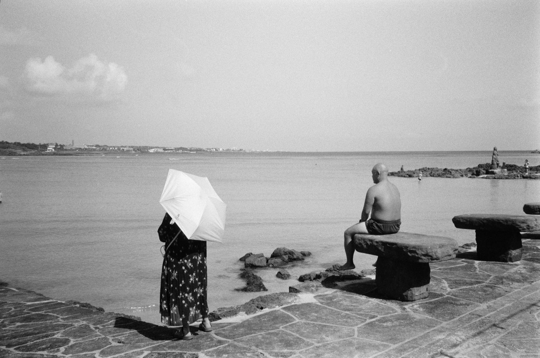 A person with a white umbrella and a seated man looking across the water, Jeju Island, 2025